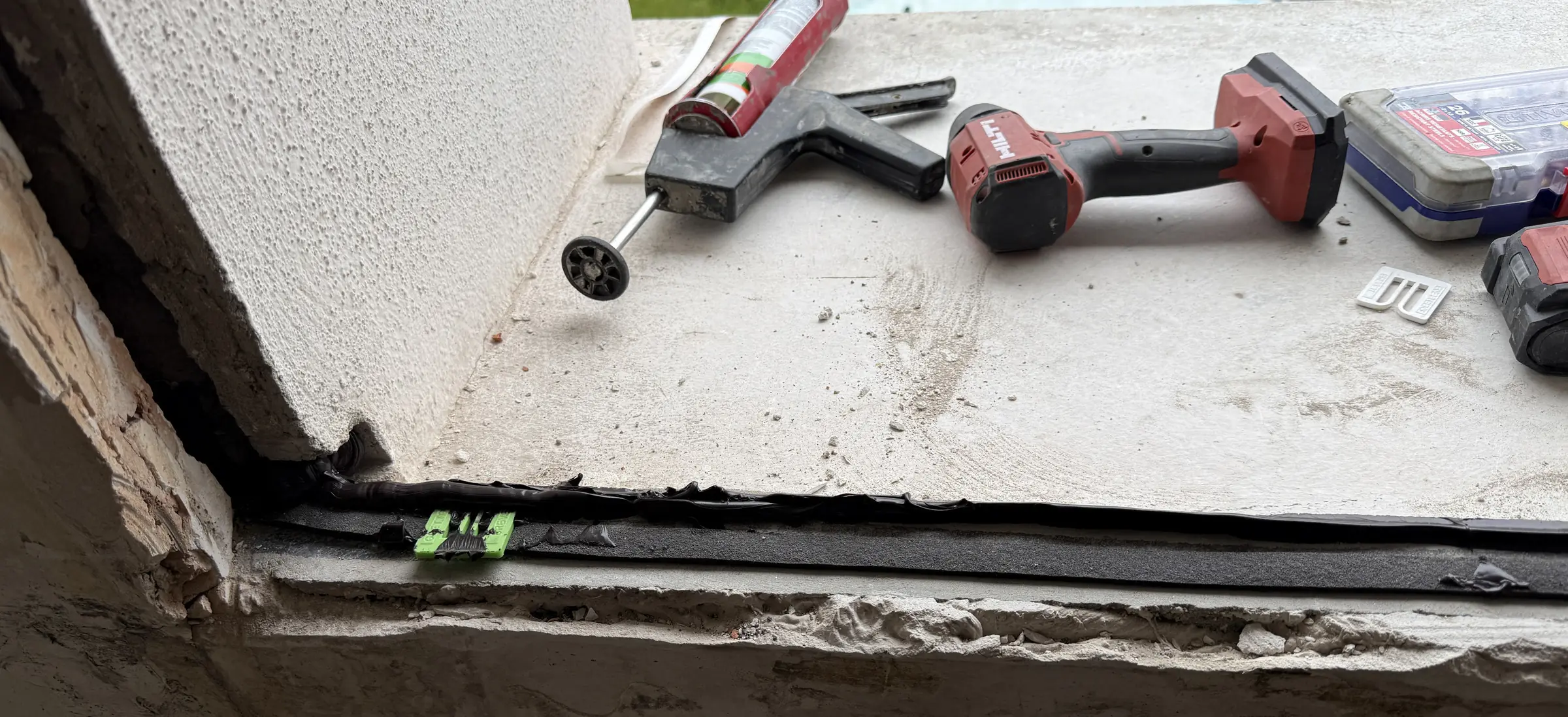 Appui de fenêtre en béton avec une bande d’étanchéité noire et des cales vertes, pistolet à mastic et visseuse posés dessus