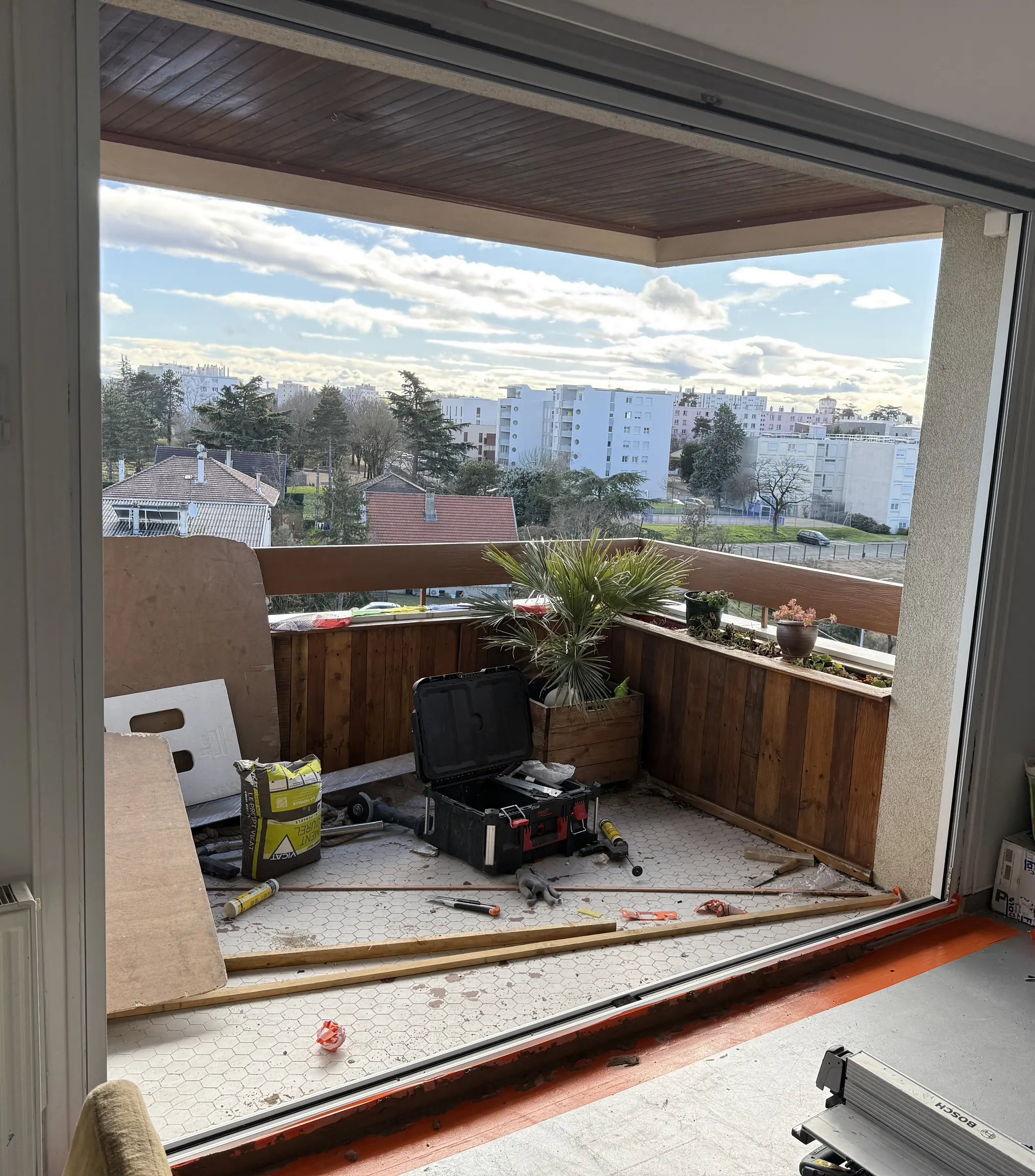 Grande ouverture vide sur un balcon après la dépose d’une baie, caisse à outils et chutes au sol, immeubles au loin