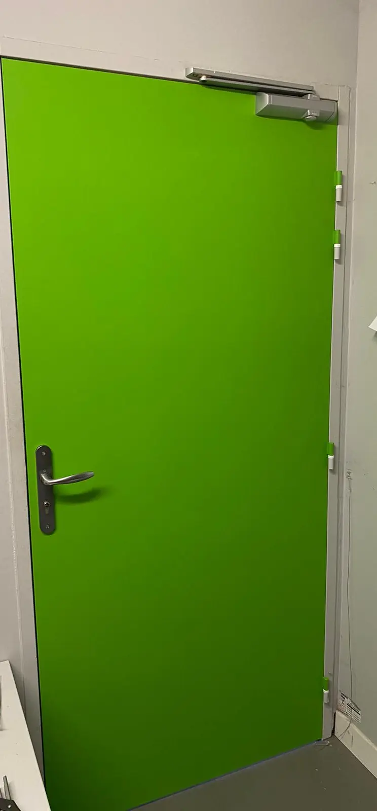 Bloc-porte métallique vert dans un local, ferme-porte en applique en haut, béquille et cylindre inox