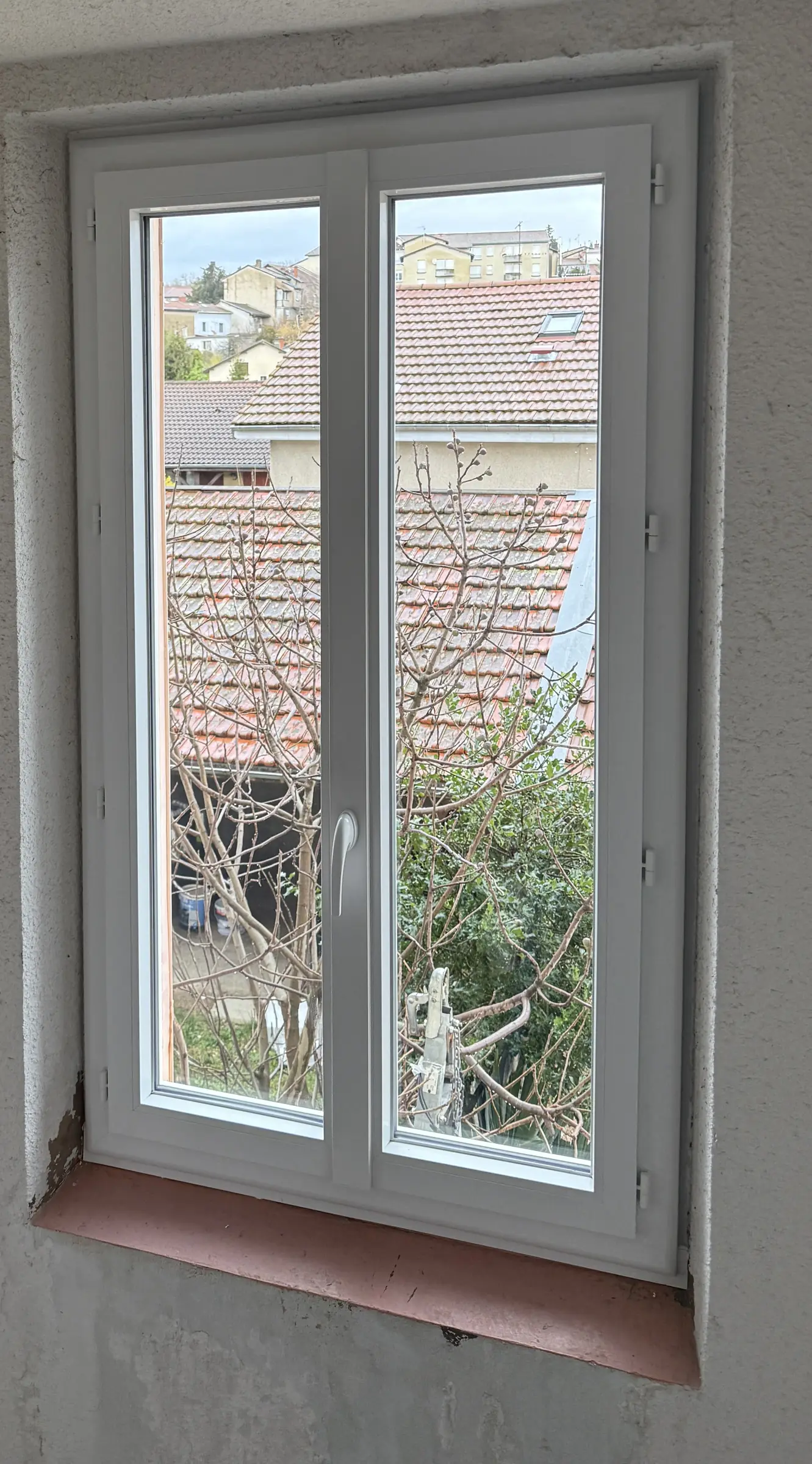 Fenêtre PVC blanche à deux vantaux dans un mur crépi blanc, appui carrelé rouge, toits de tuiles derrière