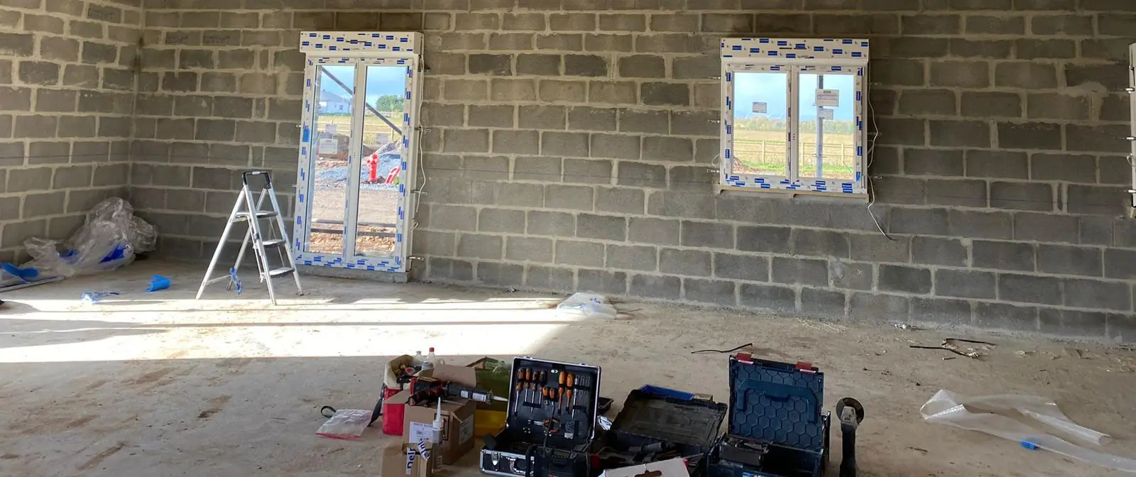 Intérieur brut d’une maison neuve : porte-fenêtre et fenêtre blanches sous films, escabeau et outillage au sol