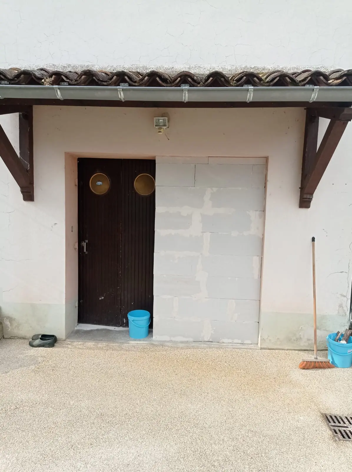 Façade sous un auvent : moitié droite de l’ouverture de garage murée en blocs de béton cellulaire, vieille porte à gauche