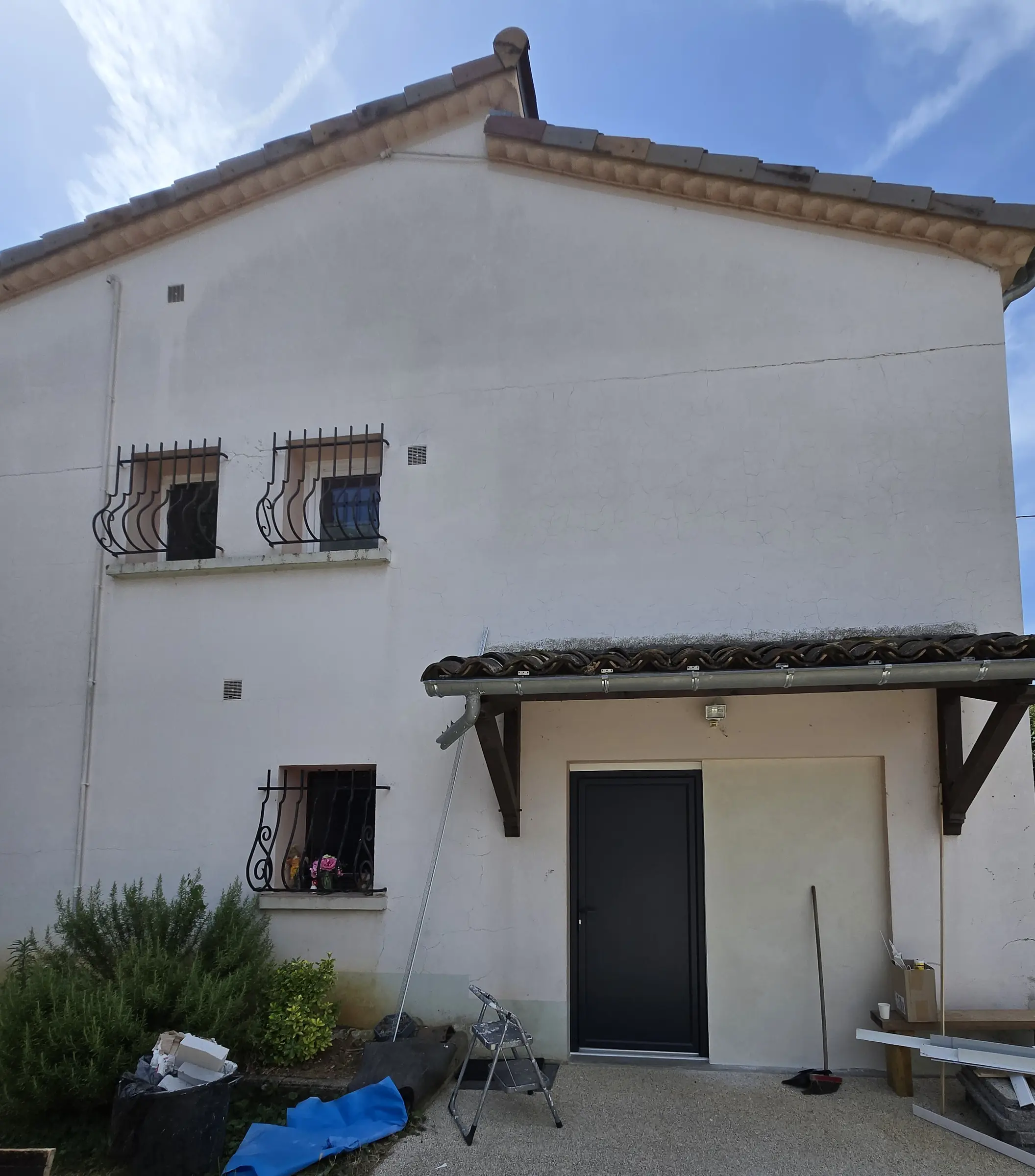 Pignon blanc d’une maison, porte de service anthracite sous un auvent, fenêtres à grilles en fer forgé