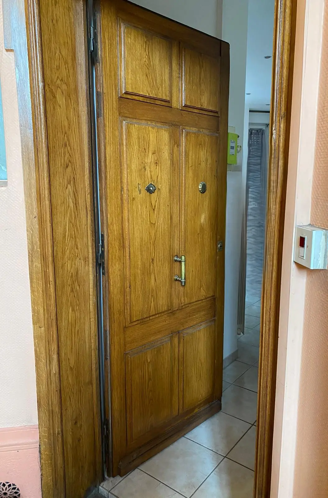 Porte d’appartement en chêne à panneaux moulurés entrouverte, couloir carrelé derrière