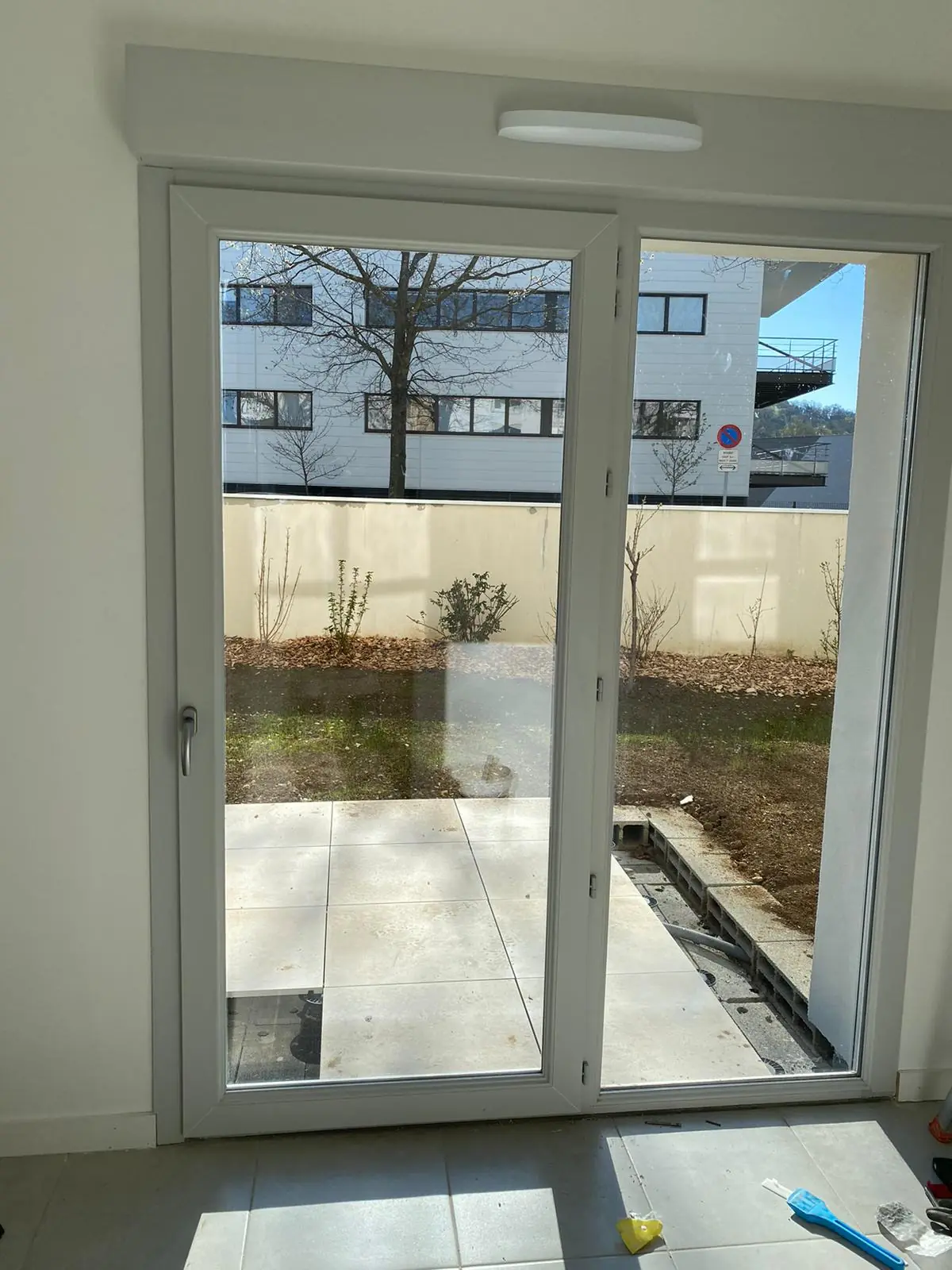 Porte-fenêtre PVC blanche à deux vantaux vue de l’intérieur, terrasse en dalles et jardin dehors