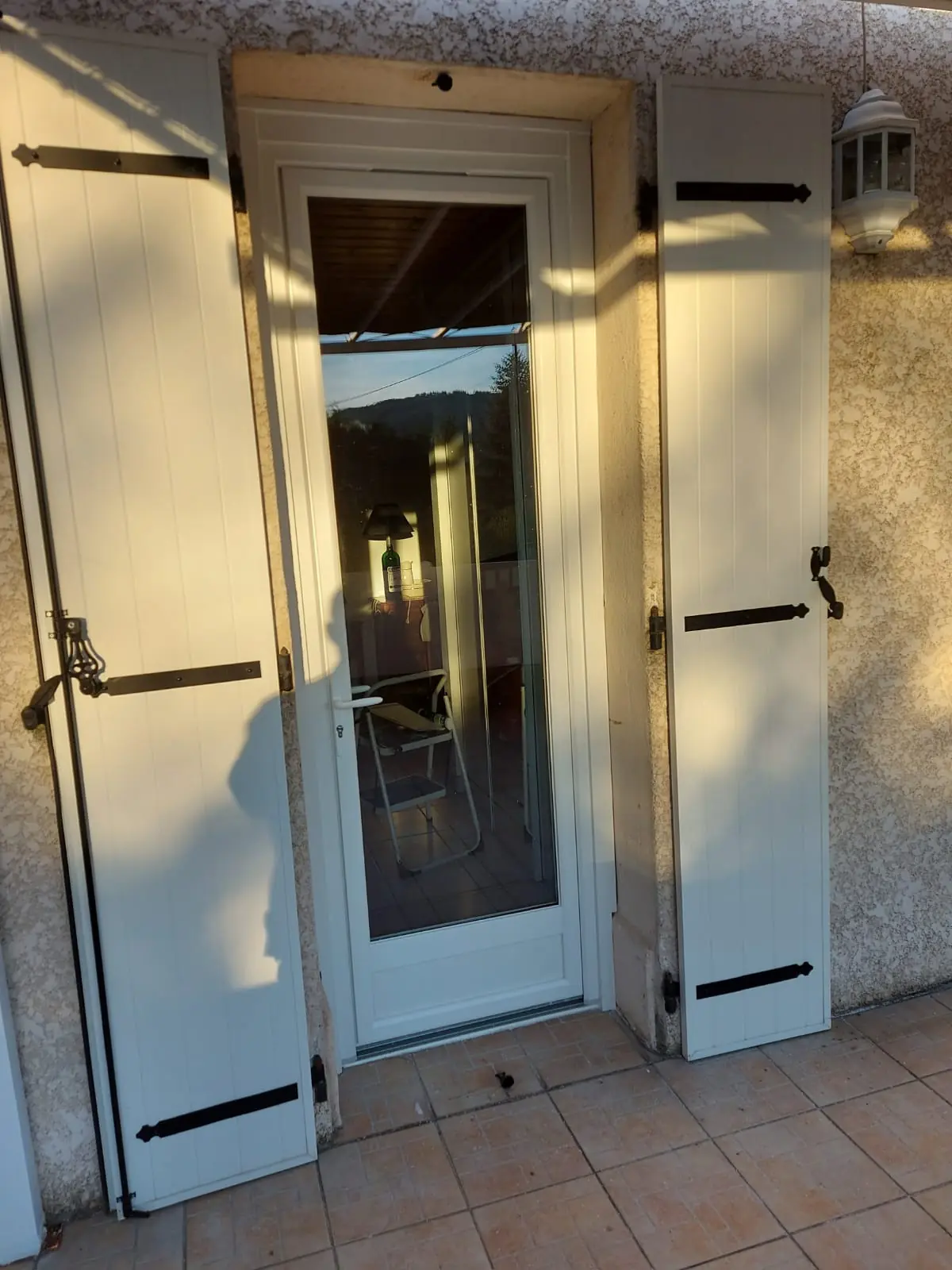 Porte-fenêtre PVC blanche vue de l’extérieur entre deux volets battants en bois blanc à pentures noires