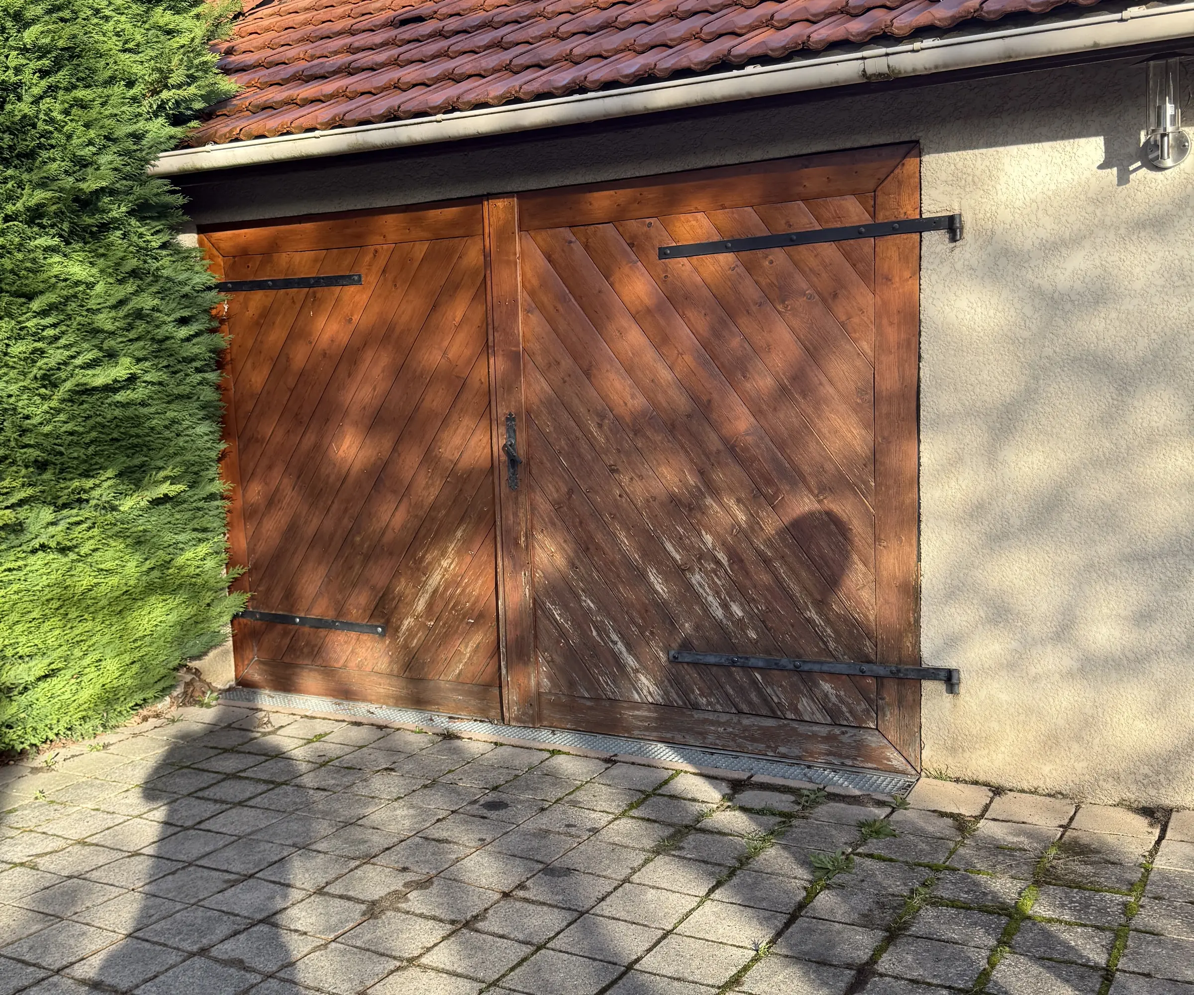 Porte de garage en bois à deux vantaux battants, lames en chevrons délavées, haie de thuyas à gauche
