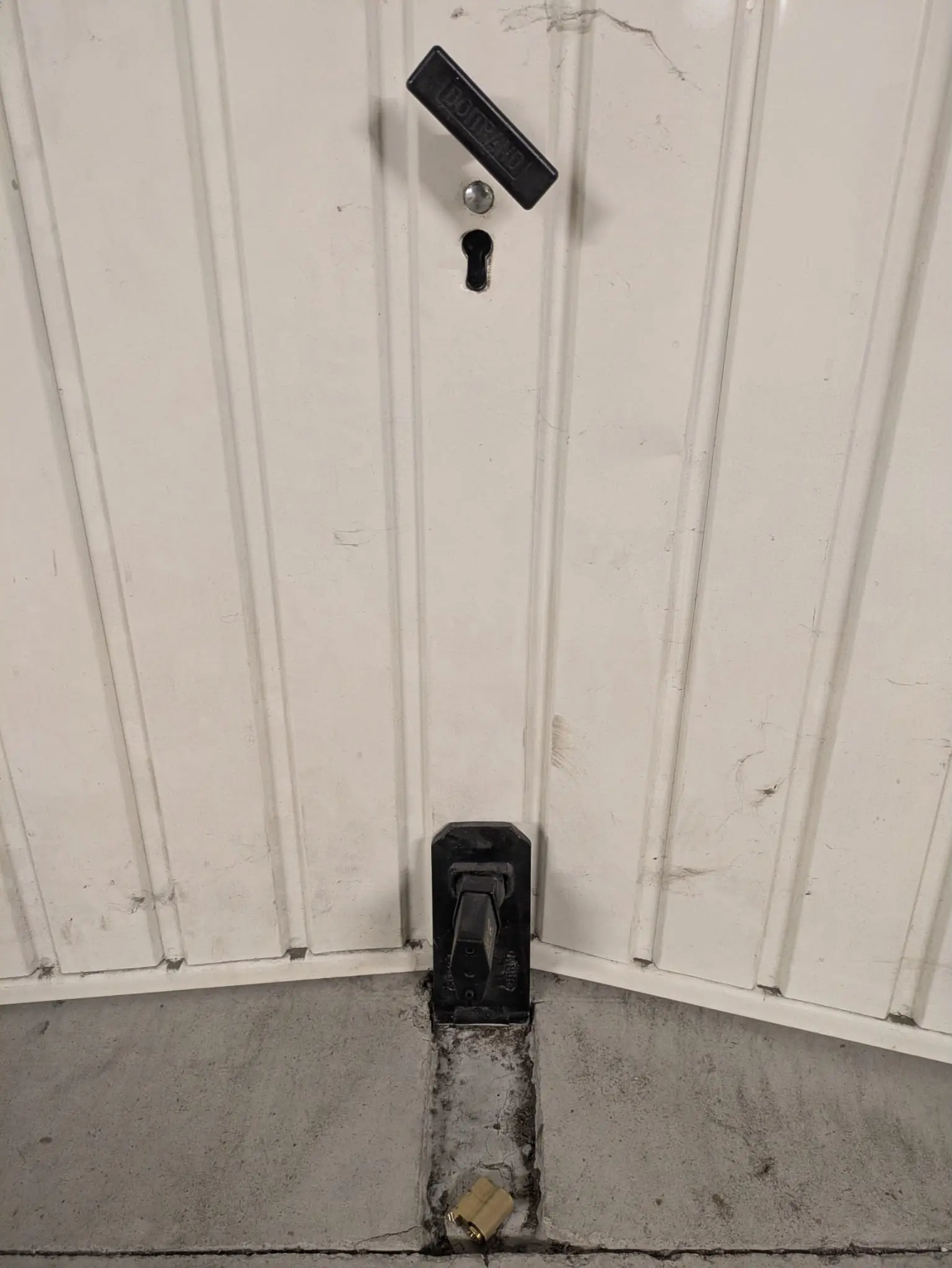 Bas d’une porte basculante blanche : poignée pendante, trou de cylindre vide, cylindre laiton tombé au sol