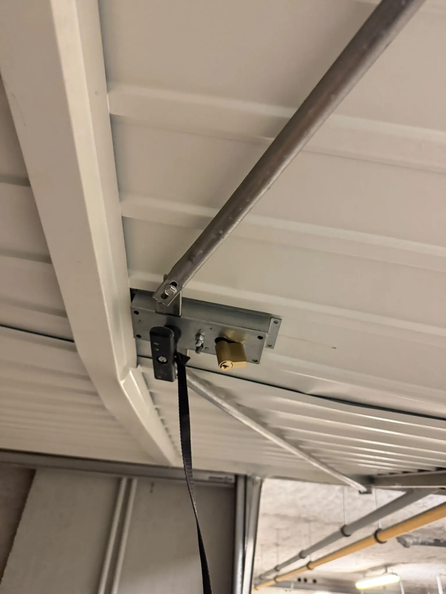 Dessous d’une porte de garage basculante ouverte : boîtier de serrure neuf, cylindre laiton et tringle de verrouillage