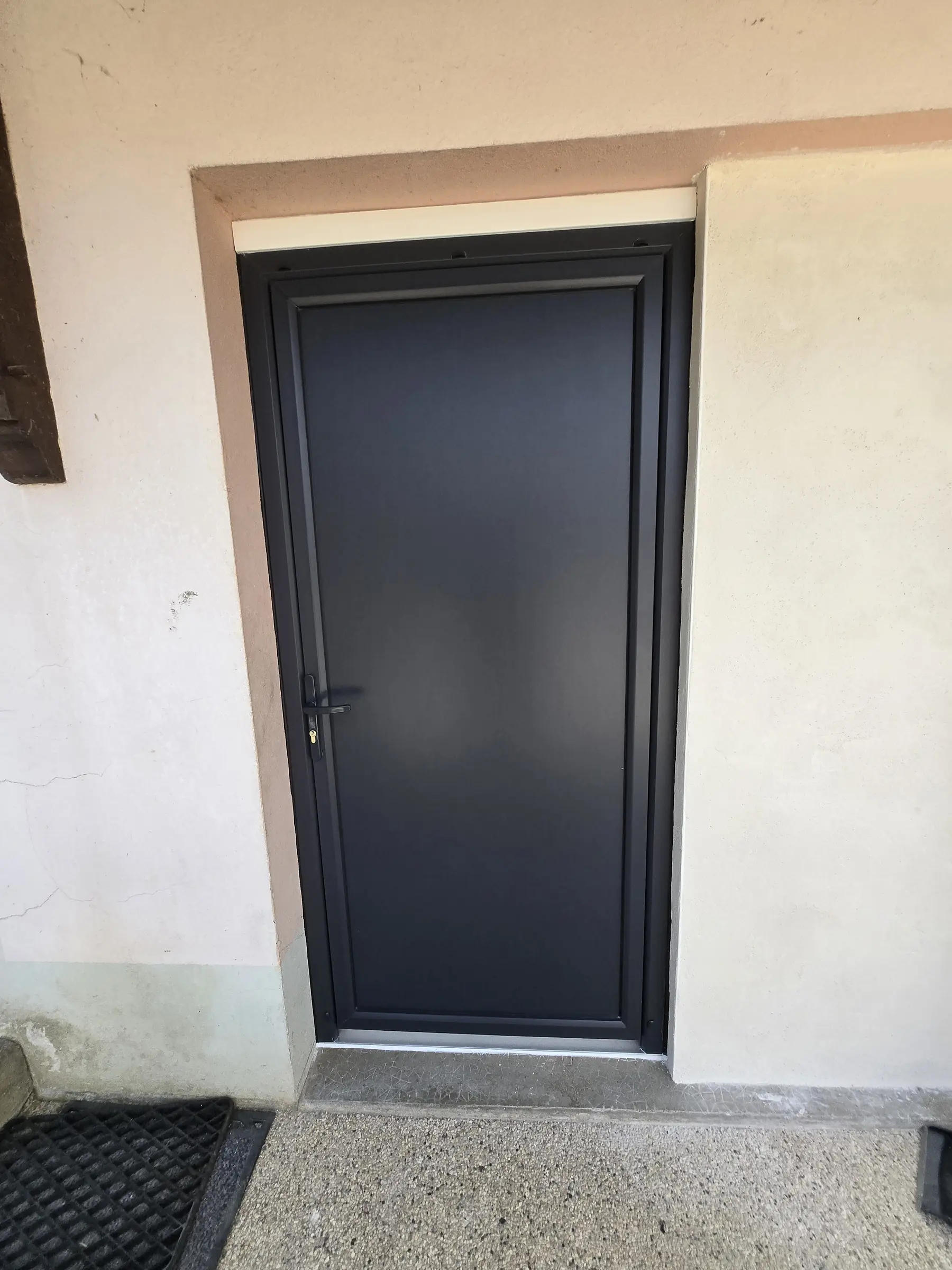Porte de service gris anthracite à panneau plein dans un encadrement crépi, partie murée enduite à droite