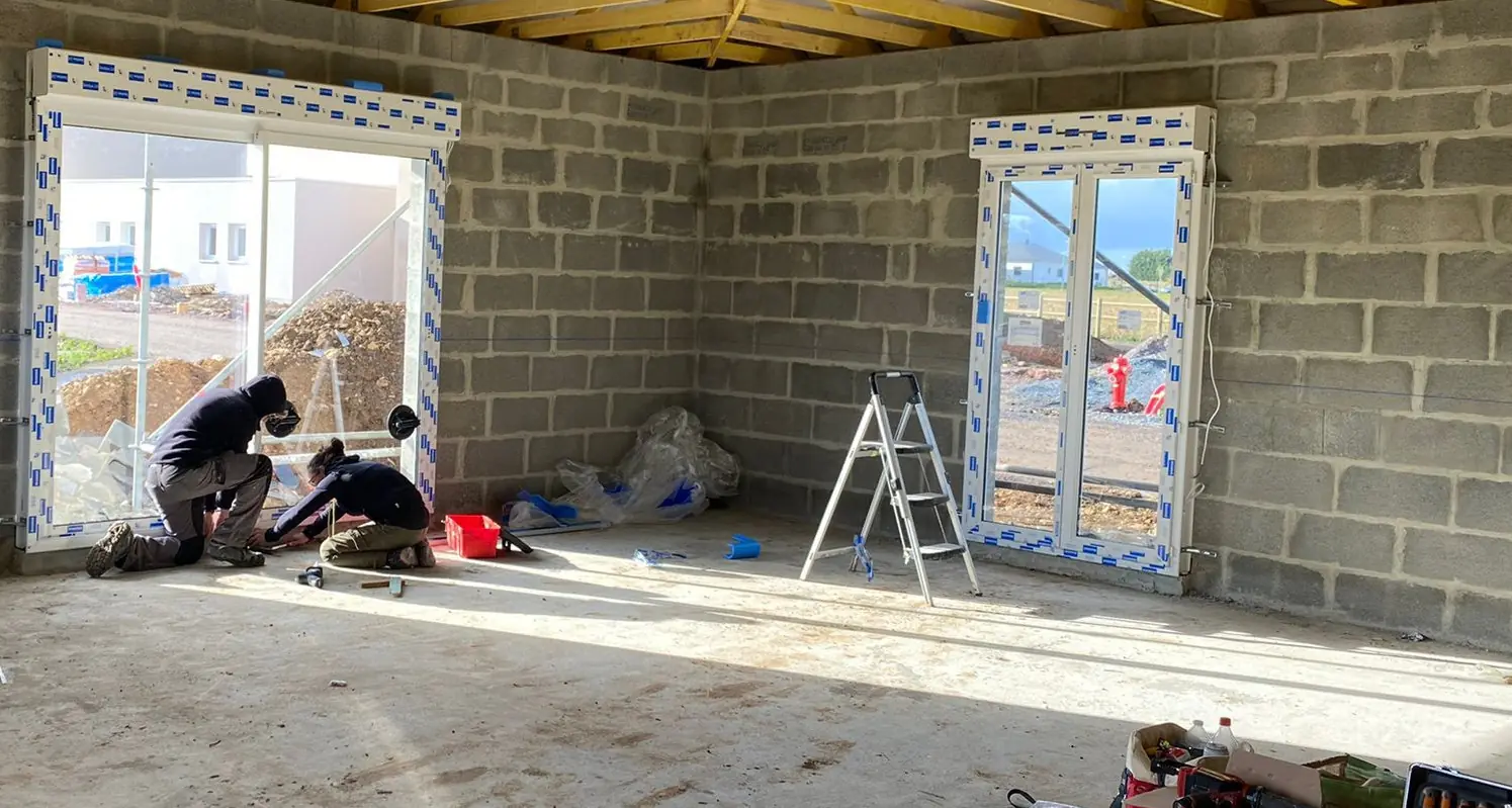 Deux poseurs penchés au pied d’une grande baie dans une maison neuve en parpaings, porte-fenêtre posée à droite